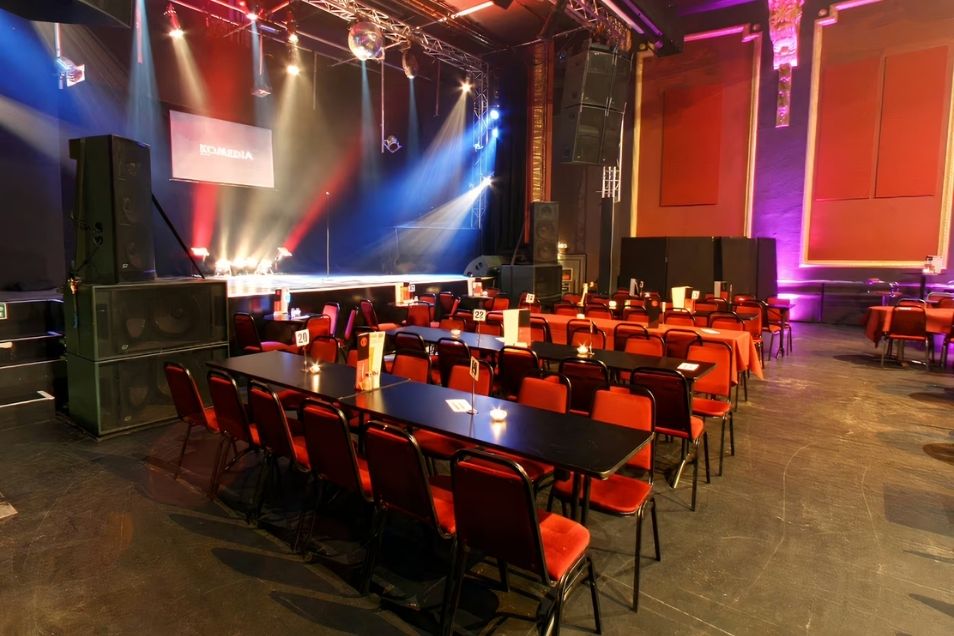 A large event space or concert hall with long tables and red chairs arranged in front of a stage with theatrical lighting.