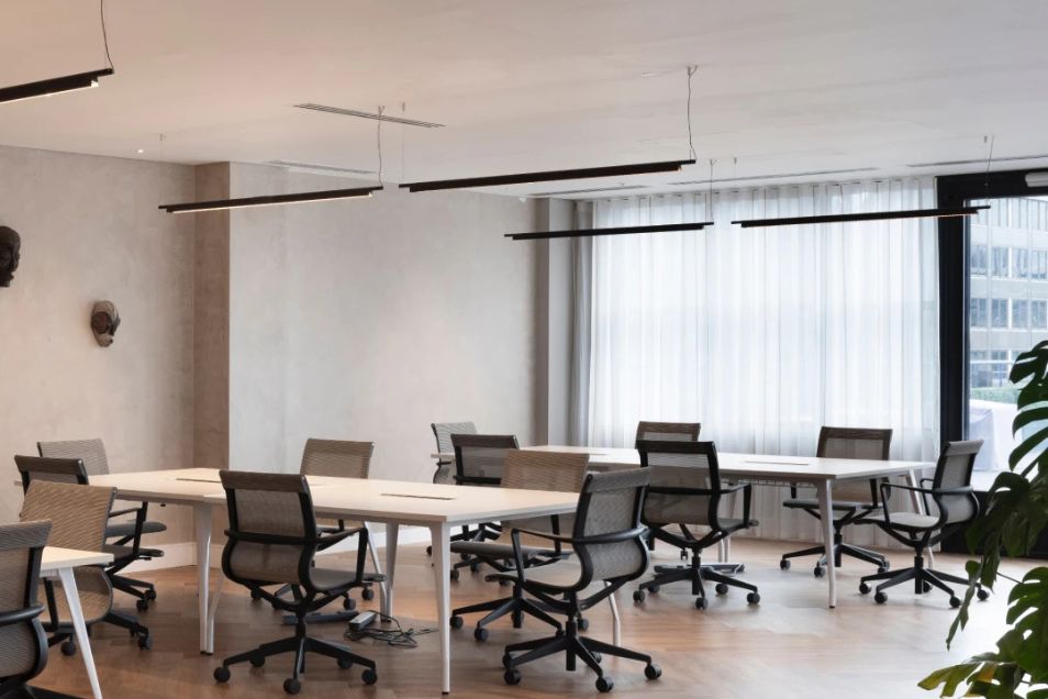 An open-plan office with white communal desks and black mesh ergonomic chairs under linear pendant lighting.