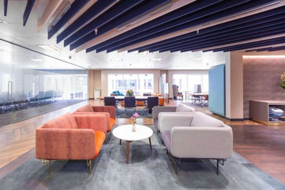 An office lounge area with coral and grey sofas arranged around a white coffee table under a slatted ceiling.