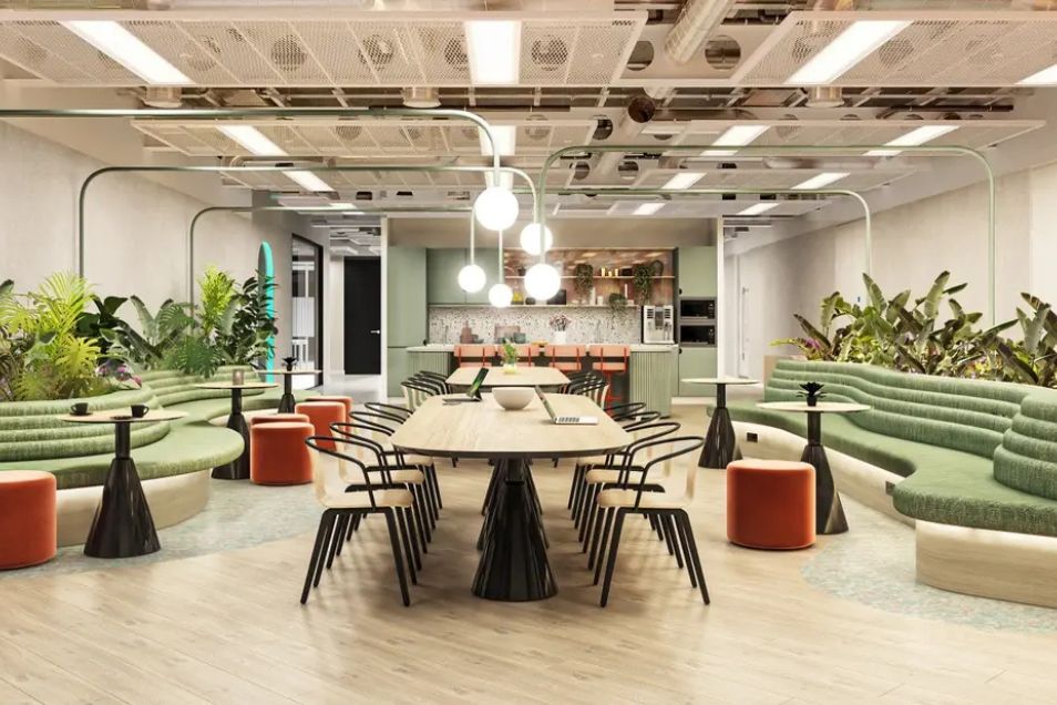 Coworking space with a long communal table, green tiered seating, and lush indoor plants under industrial lighting.