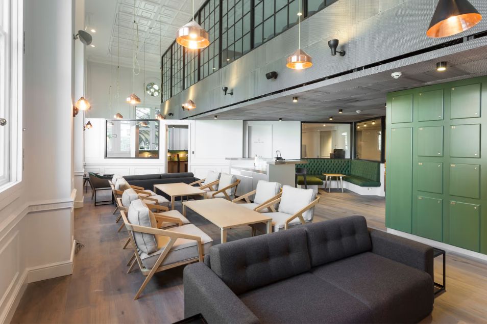 An expansive double-height office lounge with grey sofas, wooden armchairs, and a green locker wall under copper pendant lights.
