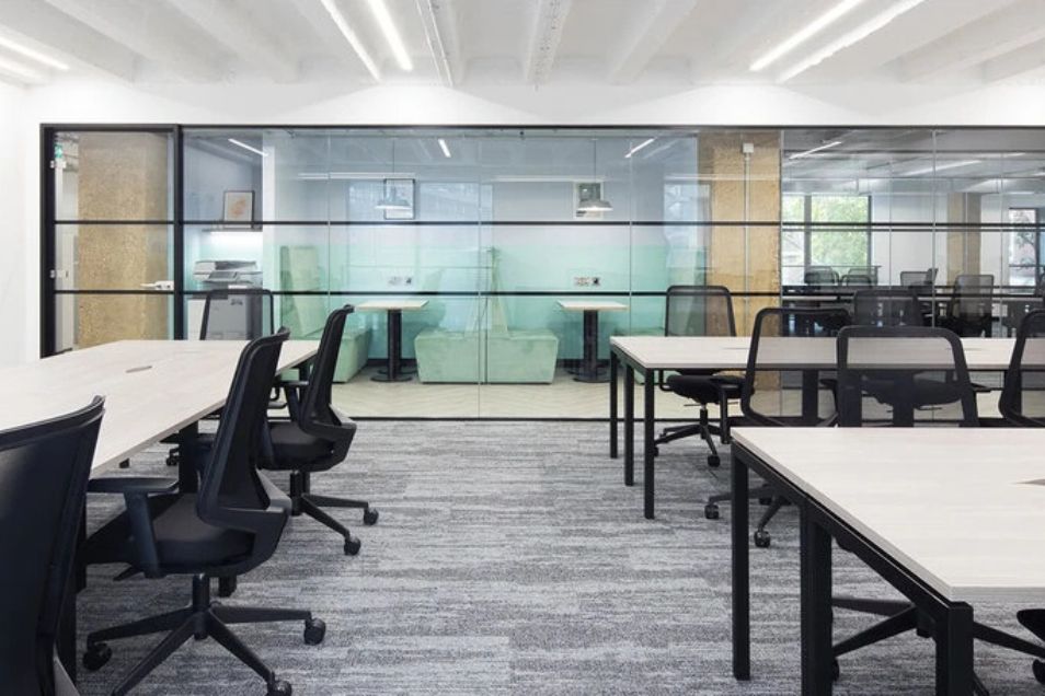 A bright, workspace featuring white desks and black chairs with a view into glass-partitioned private booths.