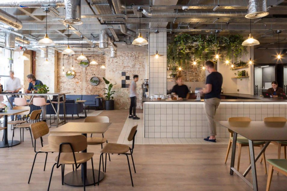 People ordering at a cafe bar in a modern office canteen with industrial lighting, wooden tables, and hanging plants.