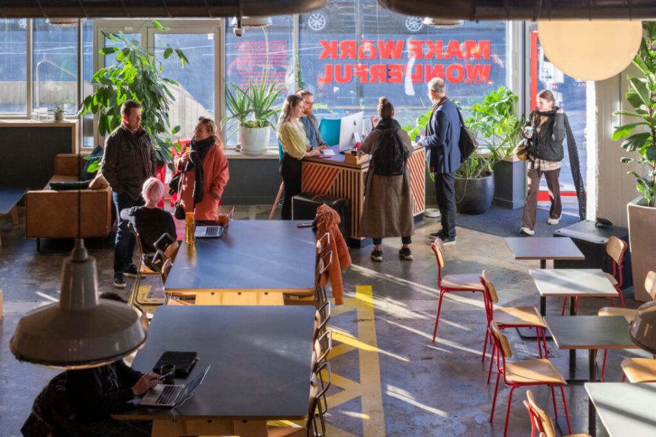 A busy office cafe where people stand and chat near a counter and sit at black tables in a sunlit space.