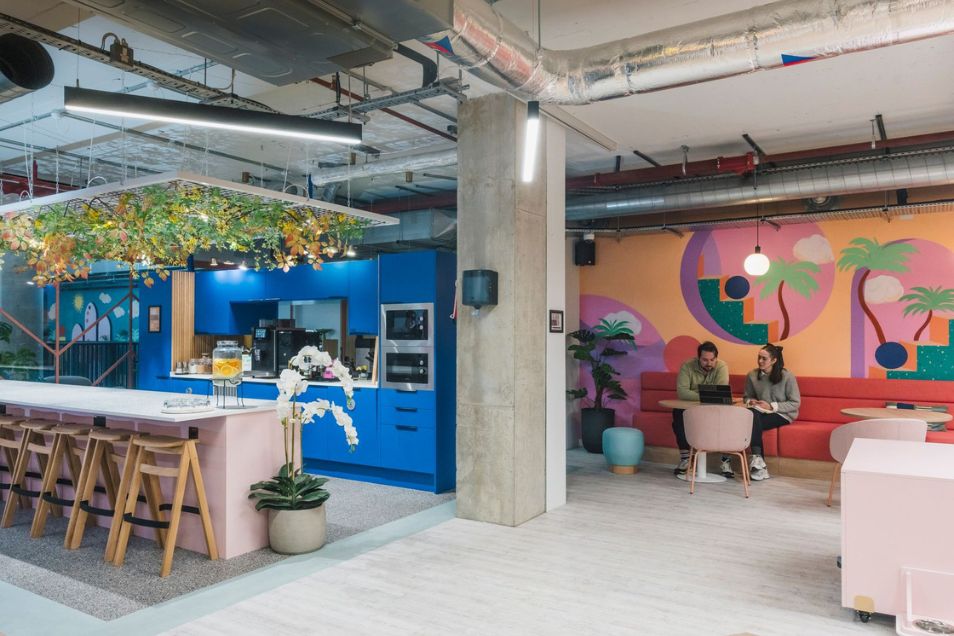Two people sit and talk in a vibrant office kitchen area featuring a pink breakfast bar and a colorful tropical mural.
