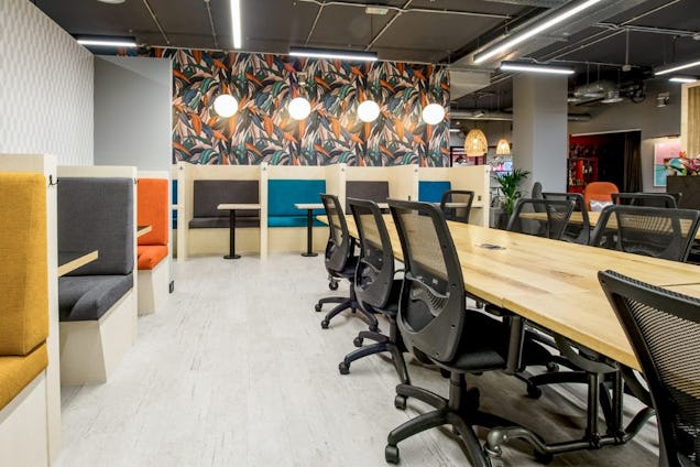 An office with long wooden communal tables, black mesh chairs, and colourful private working booths against a patterned wallpaper.