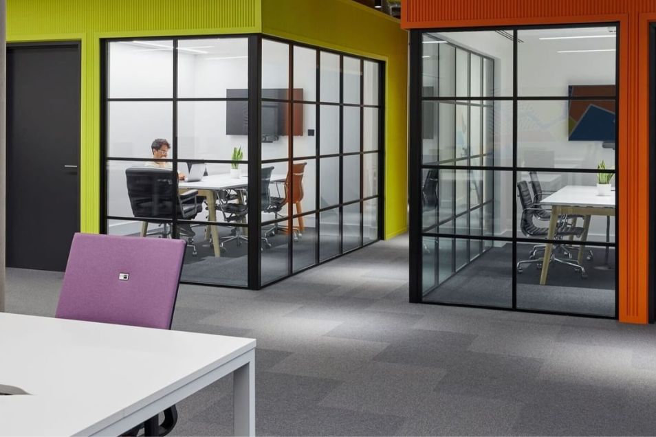 Office space with glass-partitioned meeting rooms where individuals are seated and working at white desks.