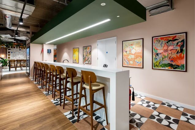 A long white breakfast bar with wooden stools set against a pale pink wall decorated with colourful framed art.