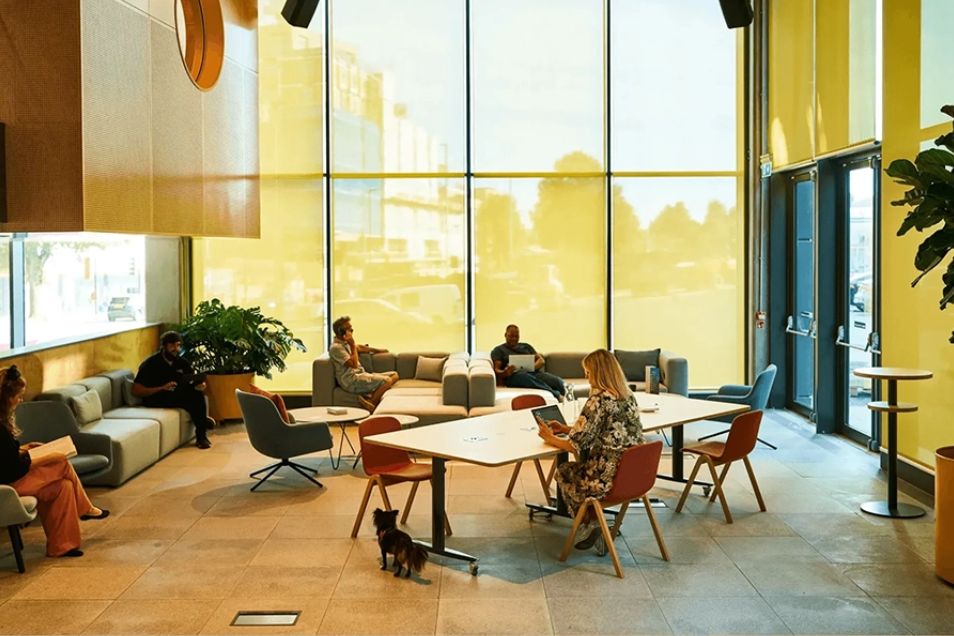 Individuals sit on sofas and at tables in a sunlit atrium with floor-to-ceiling glass windows.