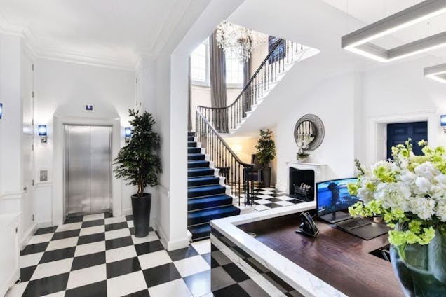 An office reception with a white staircase, a checkered floor, an elevator, and a reception desk with flowers.