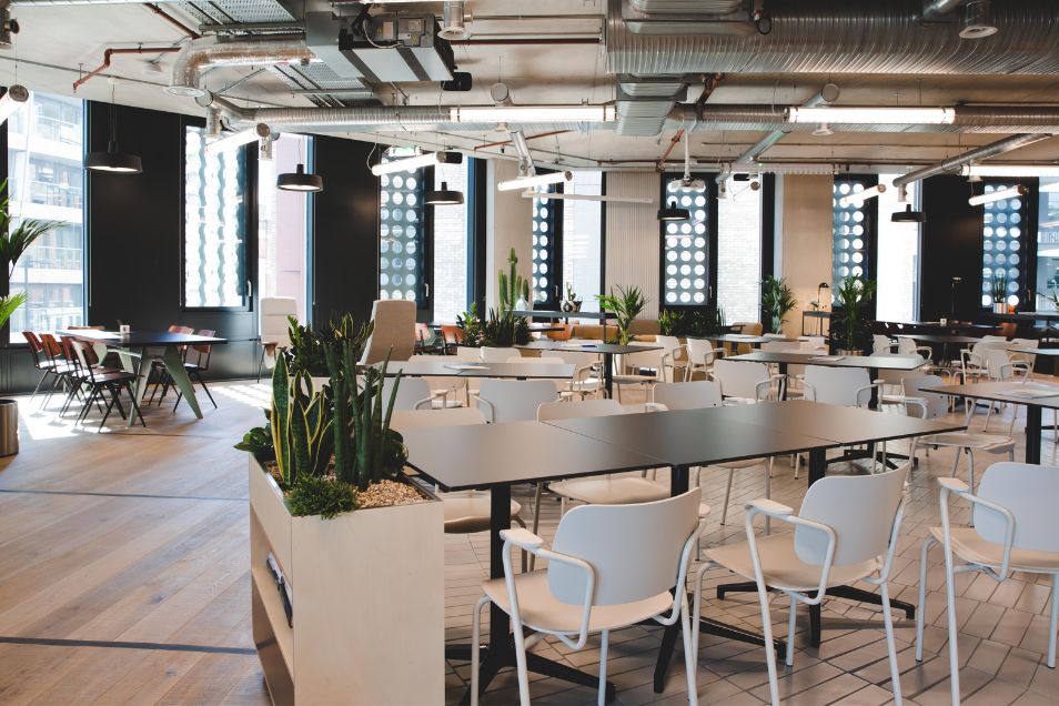 Rows of white chairs and black tables fill an office space with wooden floors and exposed ceiling pipes.
