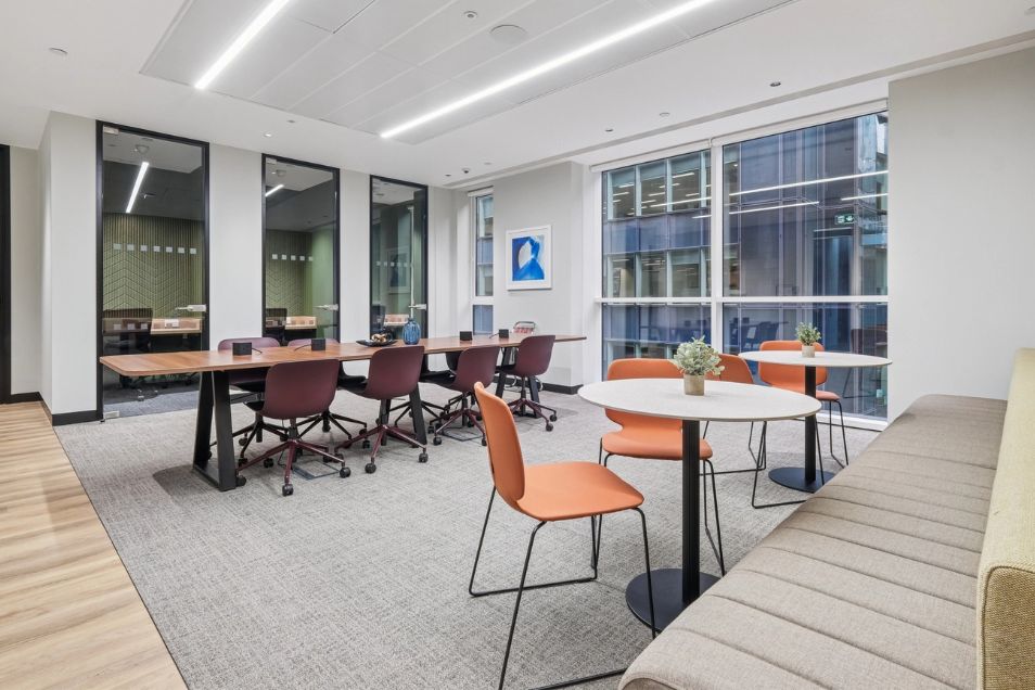 A conference room features a long table with maroon chairs and a round table with orange chairs by a window.