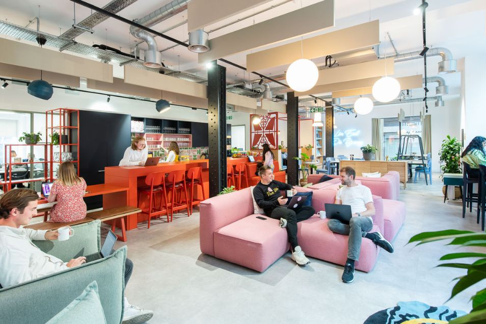 People sit on pink sofas and at a high orange table working on laptops in an open office lounge.