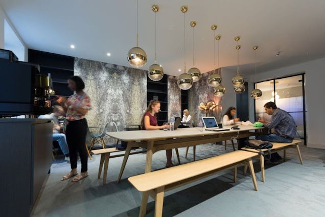 Individuals work on laptops at a wooden table with benches under several globe pendant lights in an office kitchen.