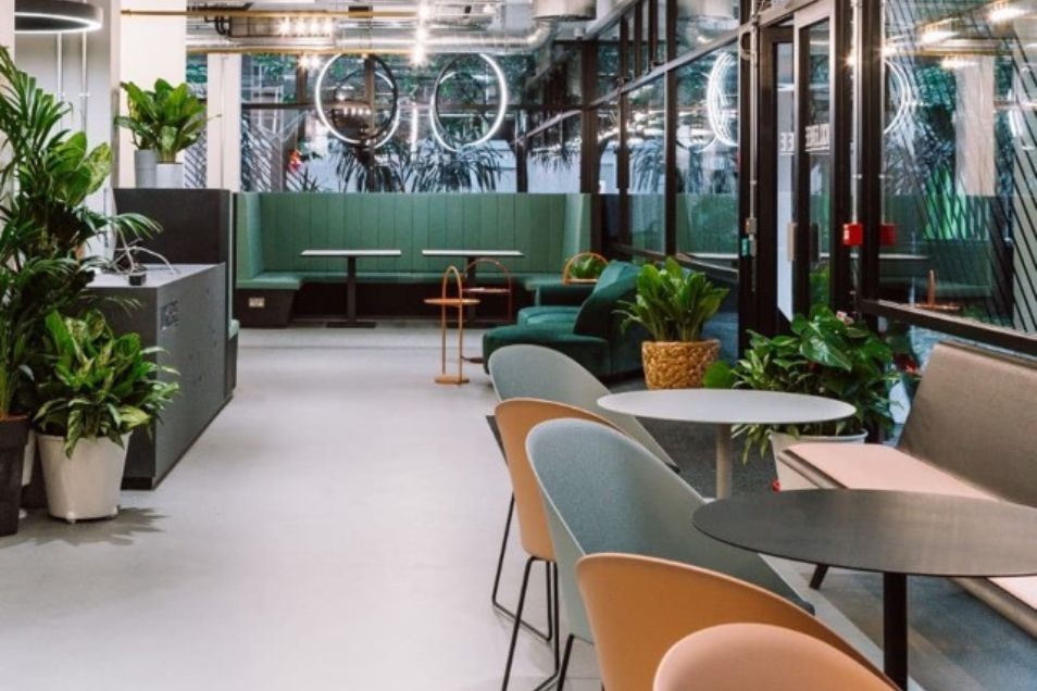 Tables and chairs are arranged in an office break area with green seating and several potted plants.
