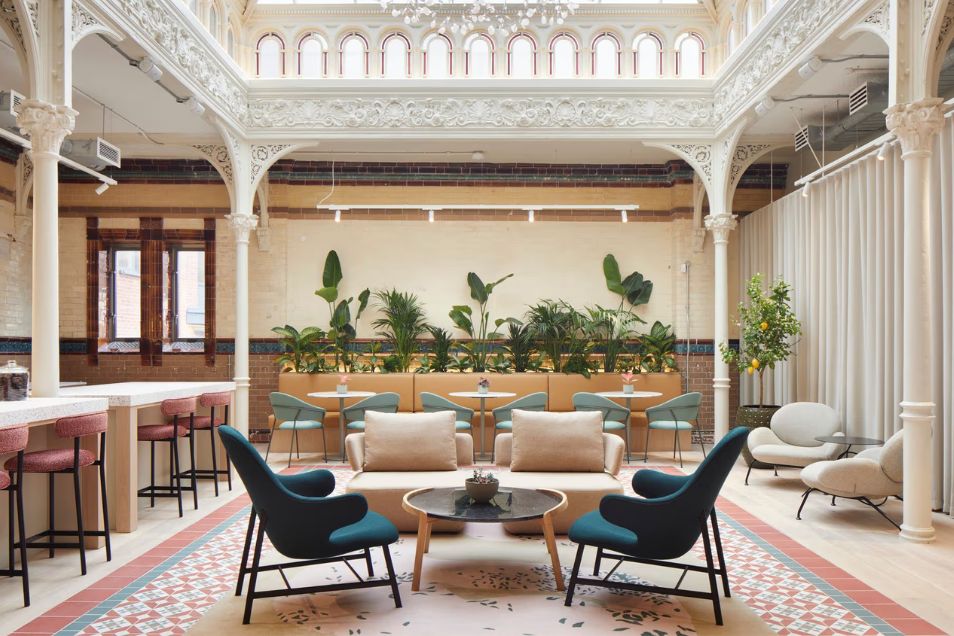 Armchairs and sofas are arranged in a lobby with pillars, plants, and a patterned rug under a skylight.
