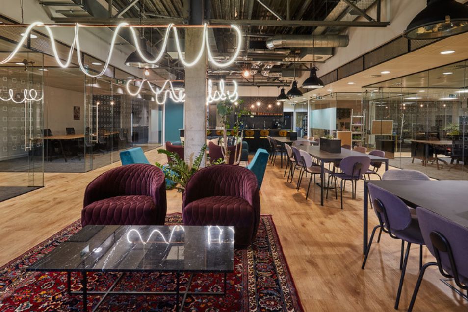 Armchairs and a coffee table on a rug in an office lounge near rows of desks and wavy ceiling lights.