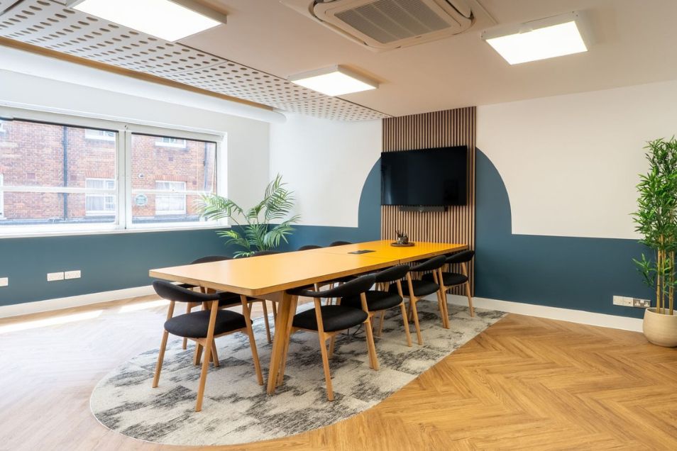 A wooden table with black chairs sits in a meeting room with a television, a rug, and a herringbone floor.