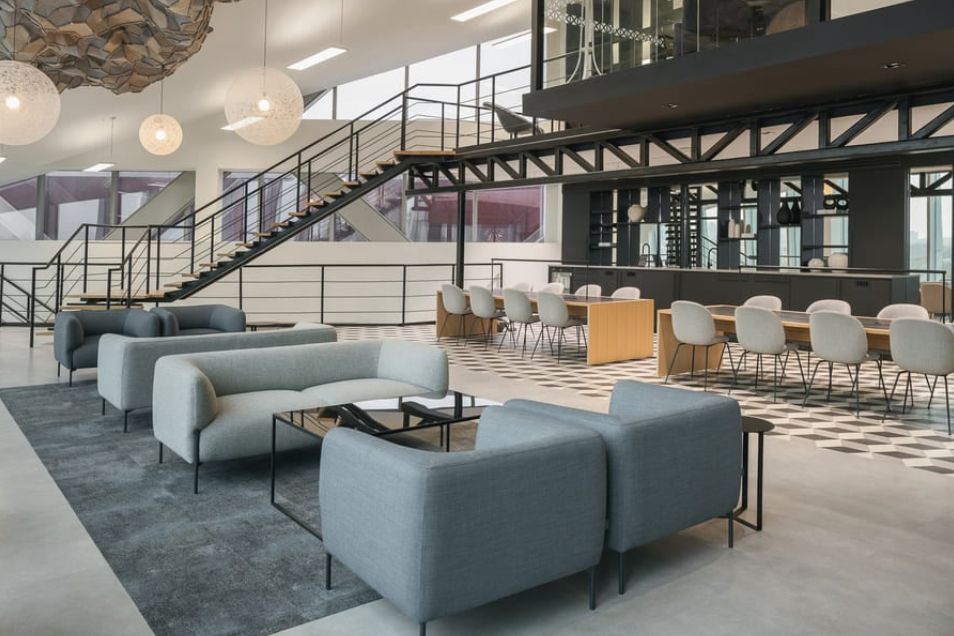 Grey sofas and long tables with chairs sit in an open office space with a staircase and mezzanine level.