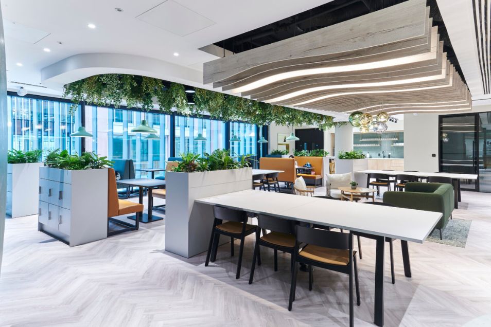 A long white table with chairs sits in a communal area with planters and a wavy ceiling feature.