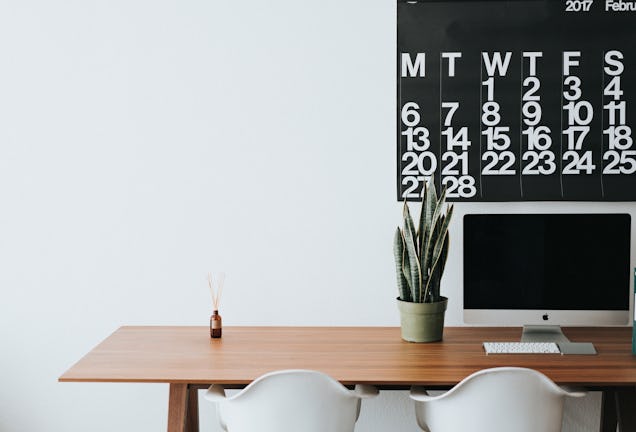 A computer, a plant, and white chairs sit at a wooden desk under a large wall calendar.