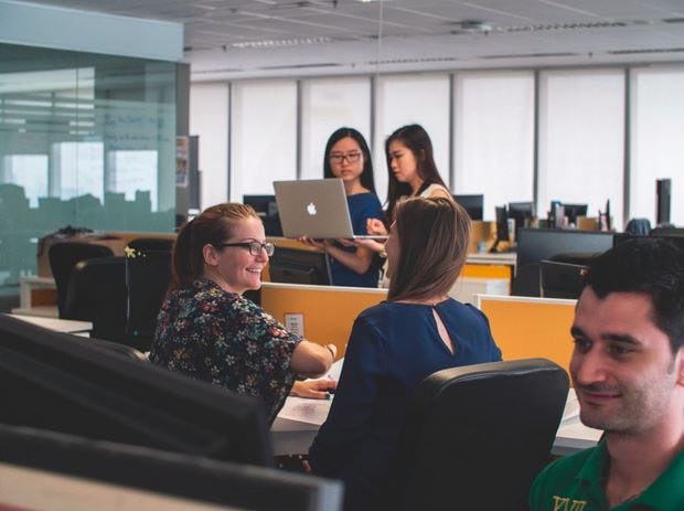 Colleagues talk and work on their laptops at a cluster of desks in an open-plan office.