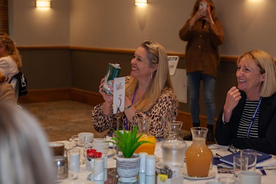 Two women sat at a table in an event space while one woman holds up a drink and smiles.