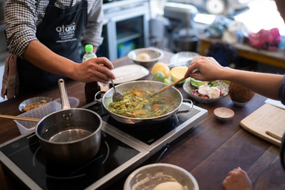 People stir a pot of food on a stovetop during a cooking lesson in a kitchen.