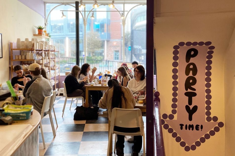 People painting at tables in a cafe next to a wall with a sign that says "PARTY TIME!".