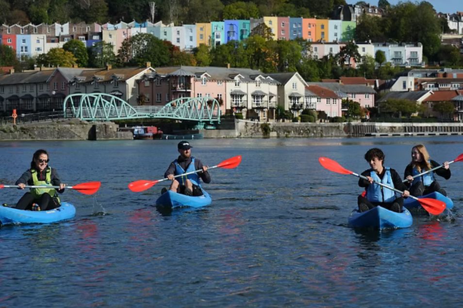 Four people paddle kayaks on a river with a bridge and a row of houses in the background.