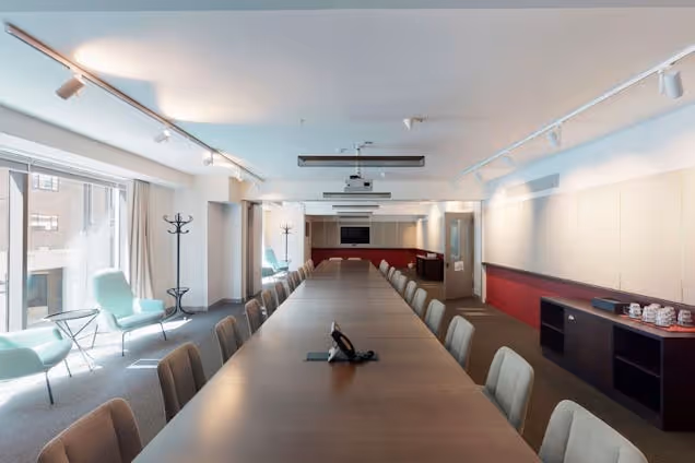 Long boardroom with a dark wood table, light grey chairs, track lighting, and a bright lounge area with mint green armchairs.