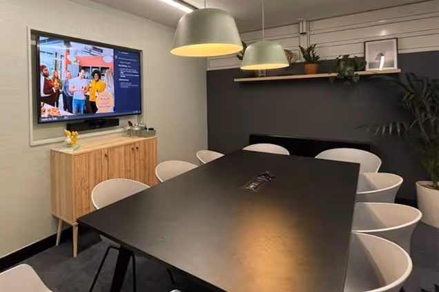 Meeting room with a black table, grey chairs, sage green pendant lights, and a wooden sideboard below a wall-mounted screen.