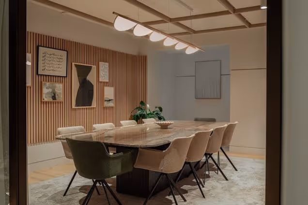 Meeting room with a stone-top table, velvet chairs, a slatted wood feature wall with framed art, and a unique wave-like pendant light.