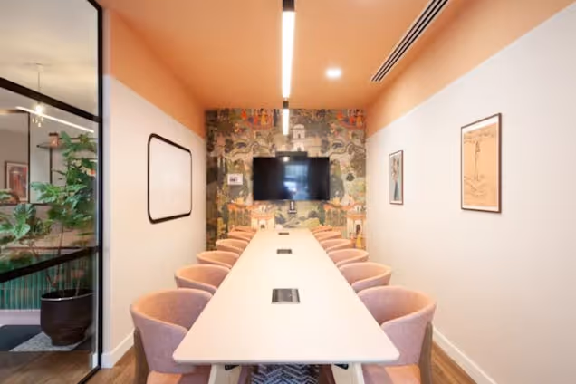 Long narrow boardroom with pink chairs, a matching peach ceiling, and an ornate landscape mural behind a wall-mounted TV.