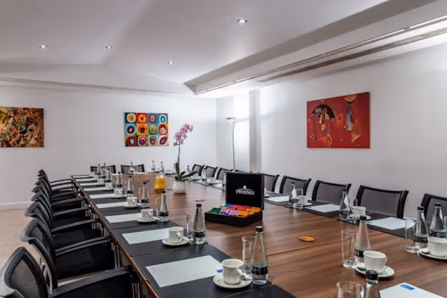 Professional boardroom featuring a long wood table set with refreshments, black mesh chairs, and vibrant abstract art on white walls.