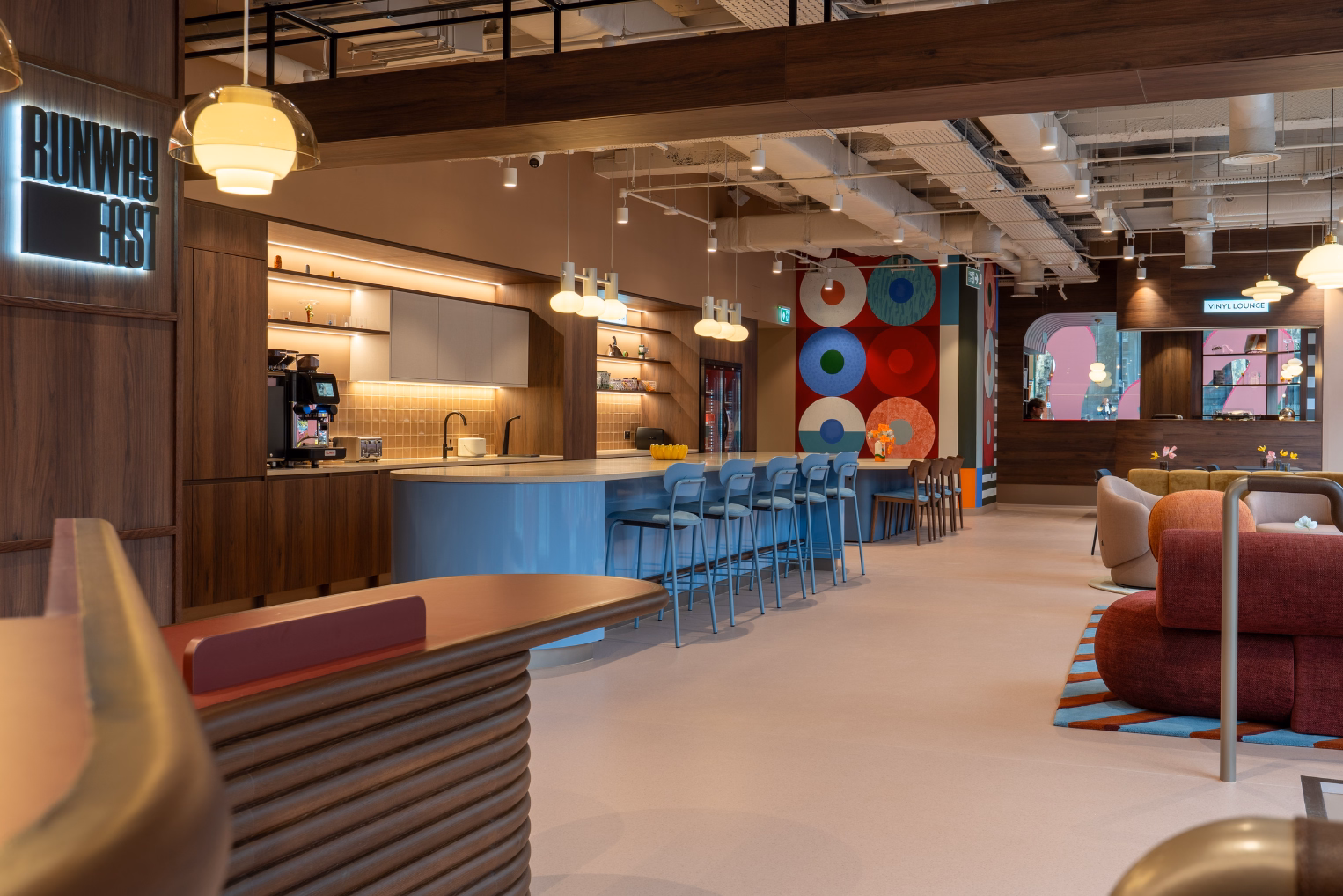 Blue stools at a blue counter in an office kitchen featuring a mural of circles, wood cabinets, burnt orange lounge seating, and lights hanging from the ceiling.
