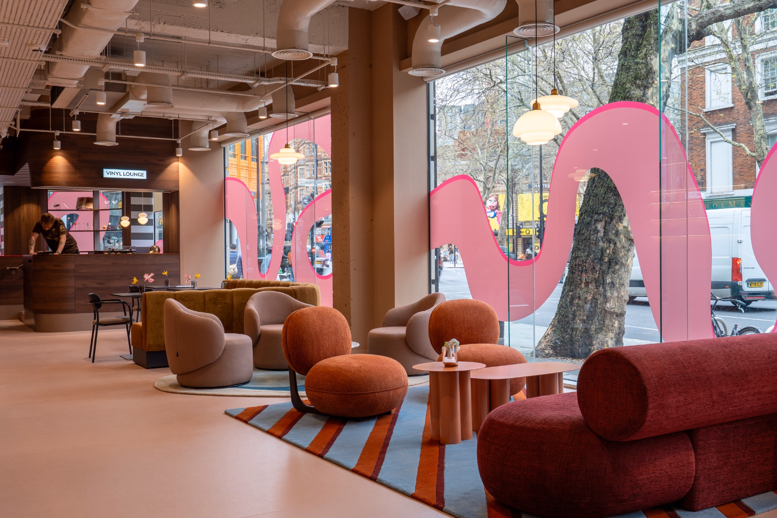 Chairs and a sofa are arranged in a lounge area next to windows with pink vinyl waves while a person works behind a wooden counter.