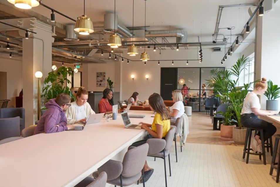 Coworking space with people working at a long pale pink table under industrial gold pendant lights and exposed ceiling pipes.