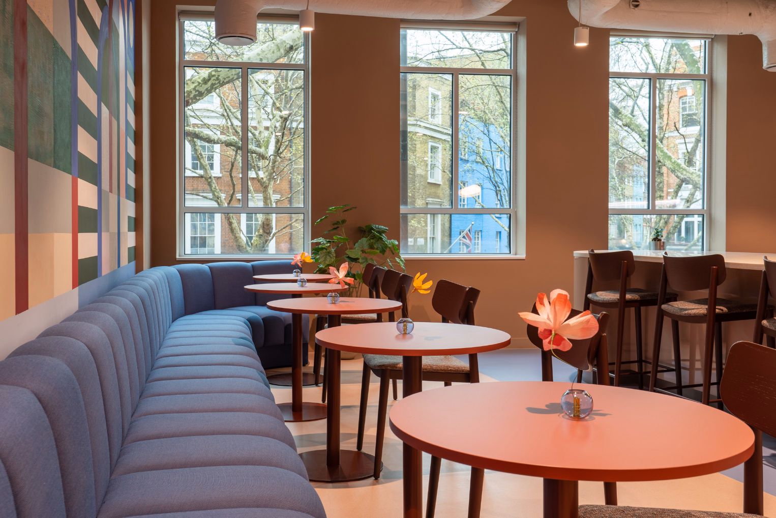 Pink tables with flowers on them in front of a blue seating space in a kitchen area featuring windows and a mural on the wall.