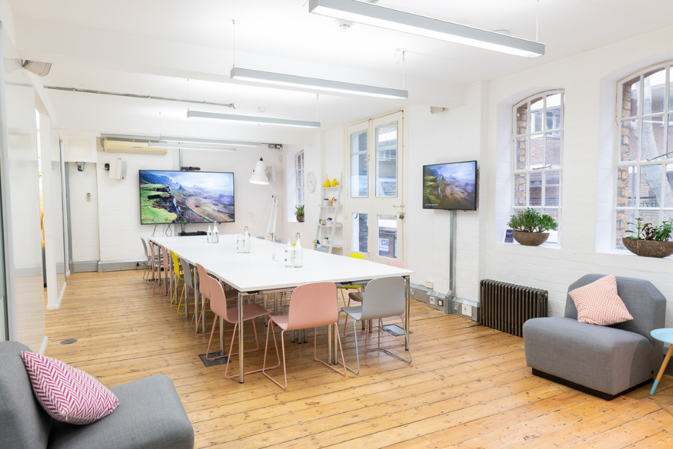 Bright, airy meeting room with a long white table, pastel chairs, pine floors, and grey armchairs with pink cushions.