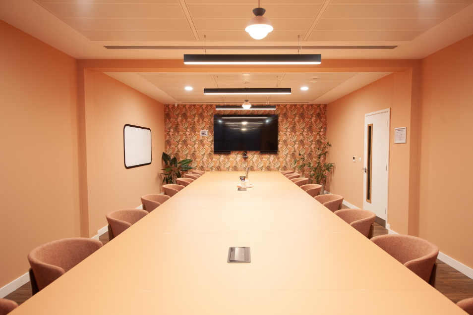 Warm-toned boardroom with a long peach table, matching curved chairs, coral walls, and a retro geometric patterned feature wall.