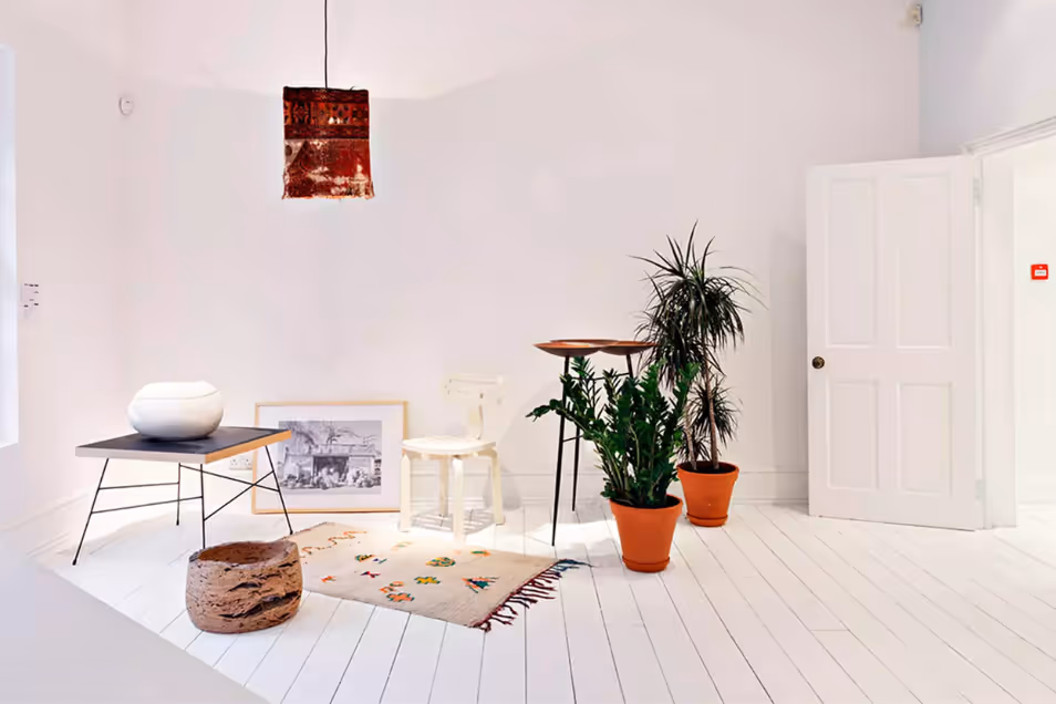 Minimalist white studio with scattered mid-century furniture, potted plants, a small patterned rug, and a copper pendant lamp.