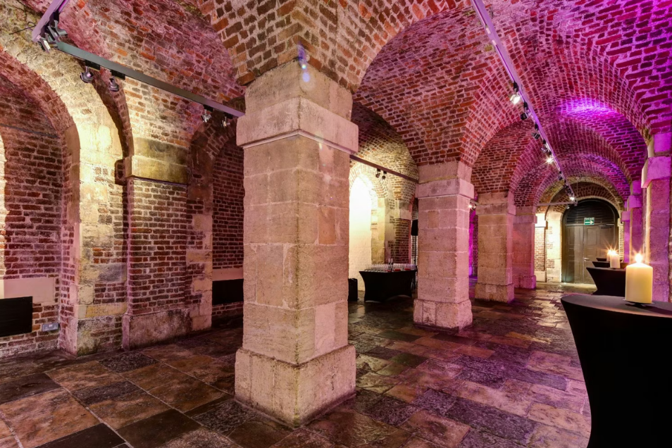 Atmospheric vaulted brick cellar with stone pillars, cobblestone floors, and corridors illuminated by purple accent lighting and candles.