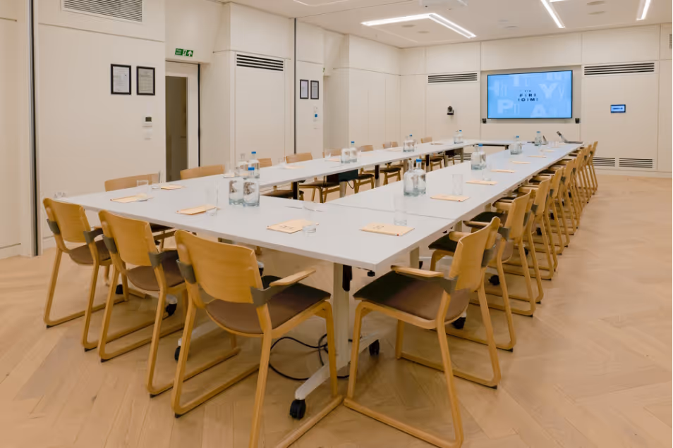 Large white u-shaped conference table surrounded by light wood chairs in a bright room with herringbone flooring.