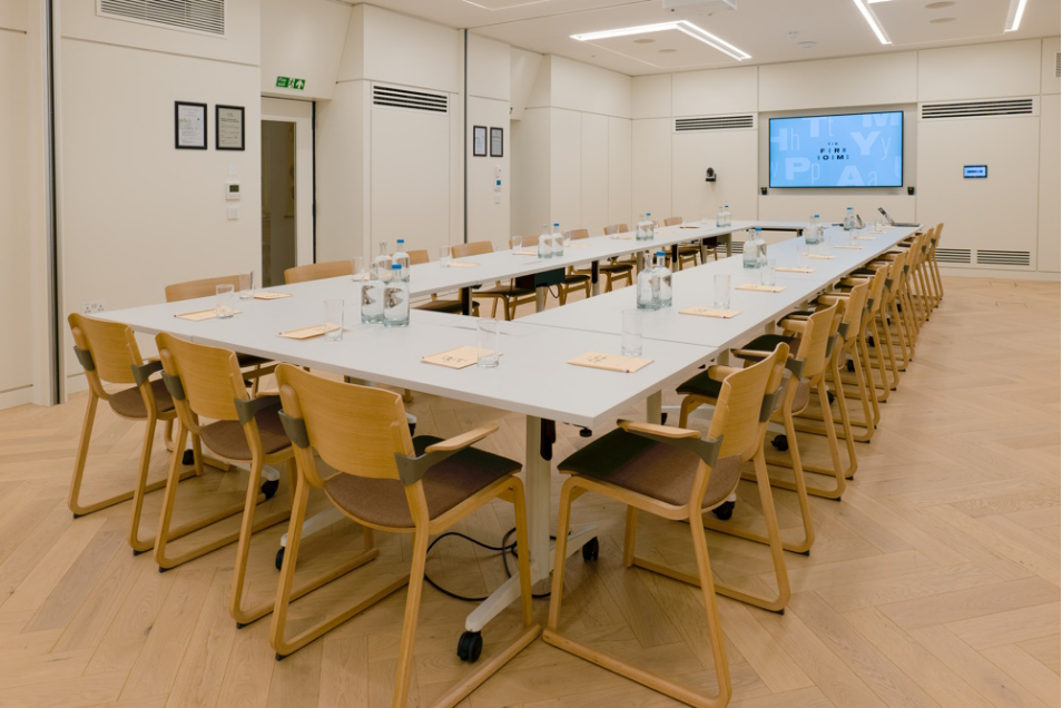 Large white u-shaped conference table surrounded by light wood chairs in a bright room with herringbone flooring.
