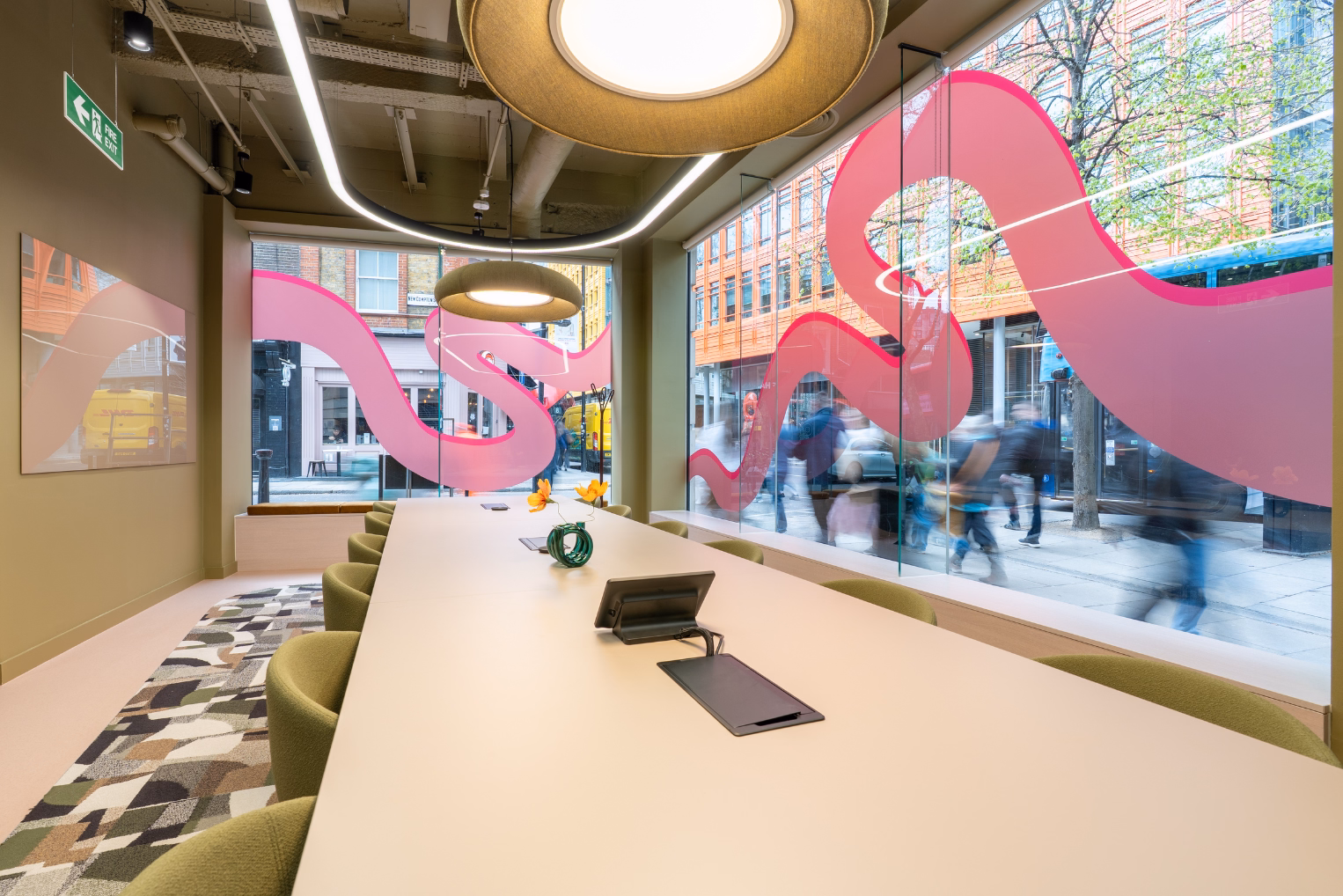 A long table with green chairs in a room with pendant lights, a patterned rug, and windows featuring pink vinyl waves as people walk by.
