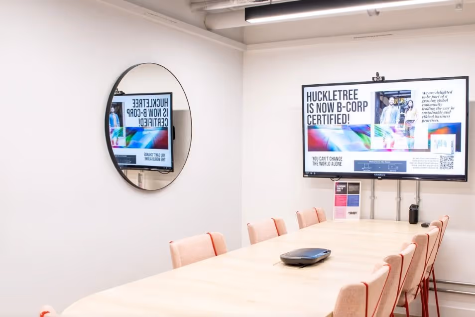 Bright meeting room with a light wood table, cream chairs with red accents, a large wall-mounted screen, and a circular mirror.