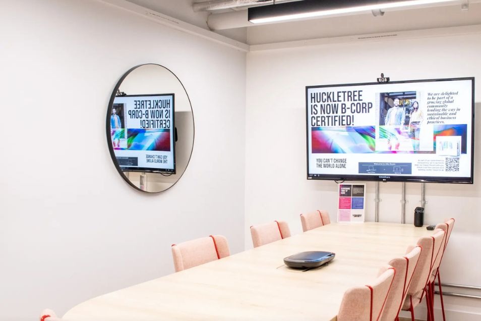 Bright meeting room with a light wood table, cream chairs with red accents, a large wall-mounted screen, and a circular mirror.