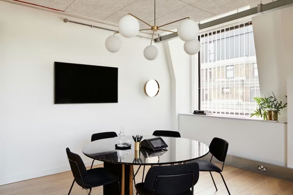 Meeting room with a round black table, matching chairs, a large globe chandelier, and a wall-mounted flat screen.