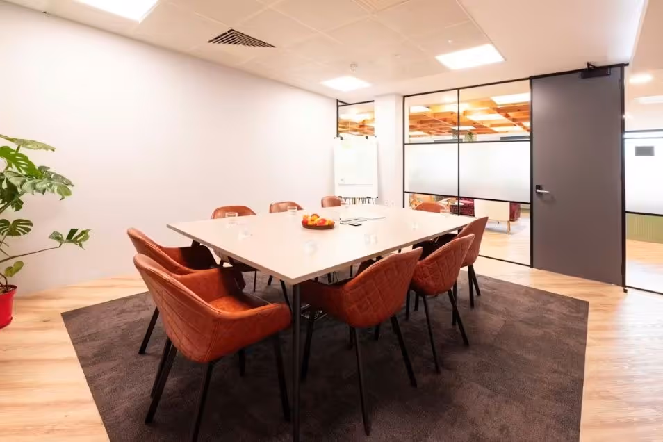 Meeting room with a light square table, rust-coloured quilted chairs, a dark area rug, and wood-look flooring.